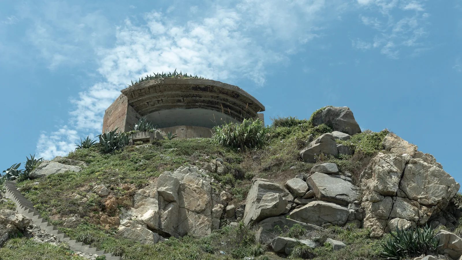 岩石與草木覆蓋的坡頂上，一座弧形混凝土建物面向鏡頭，後方是藍天與白雲。