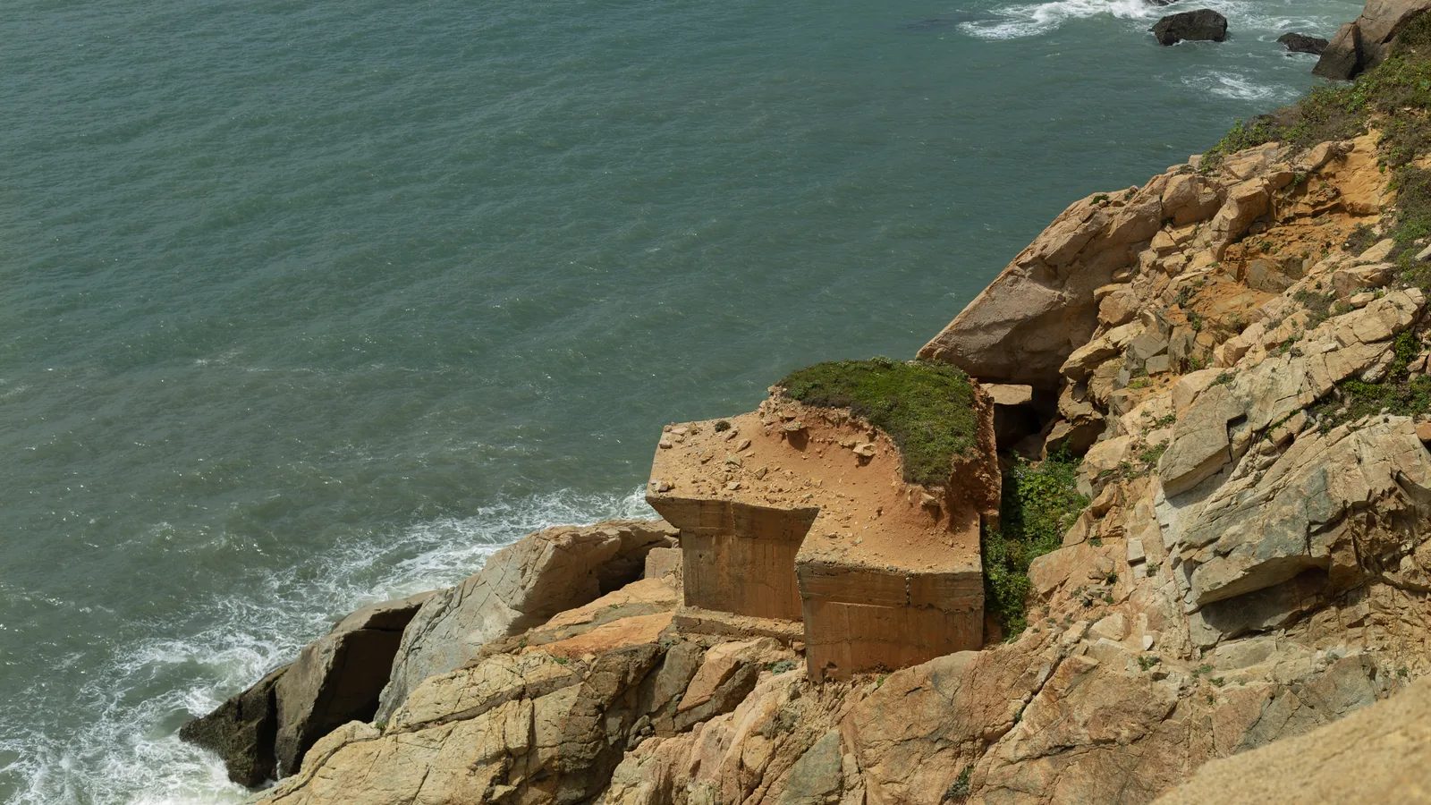 土黃色的低矮建物嵌在海邊岩坡之間，海浪沿著下方岸邊形成白色水痕。