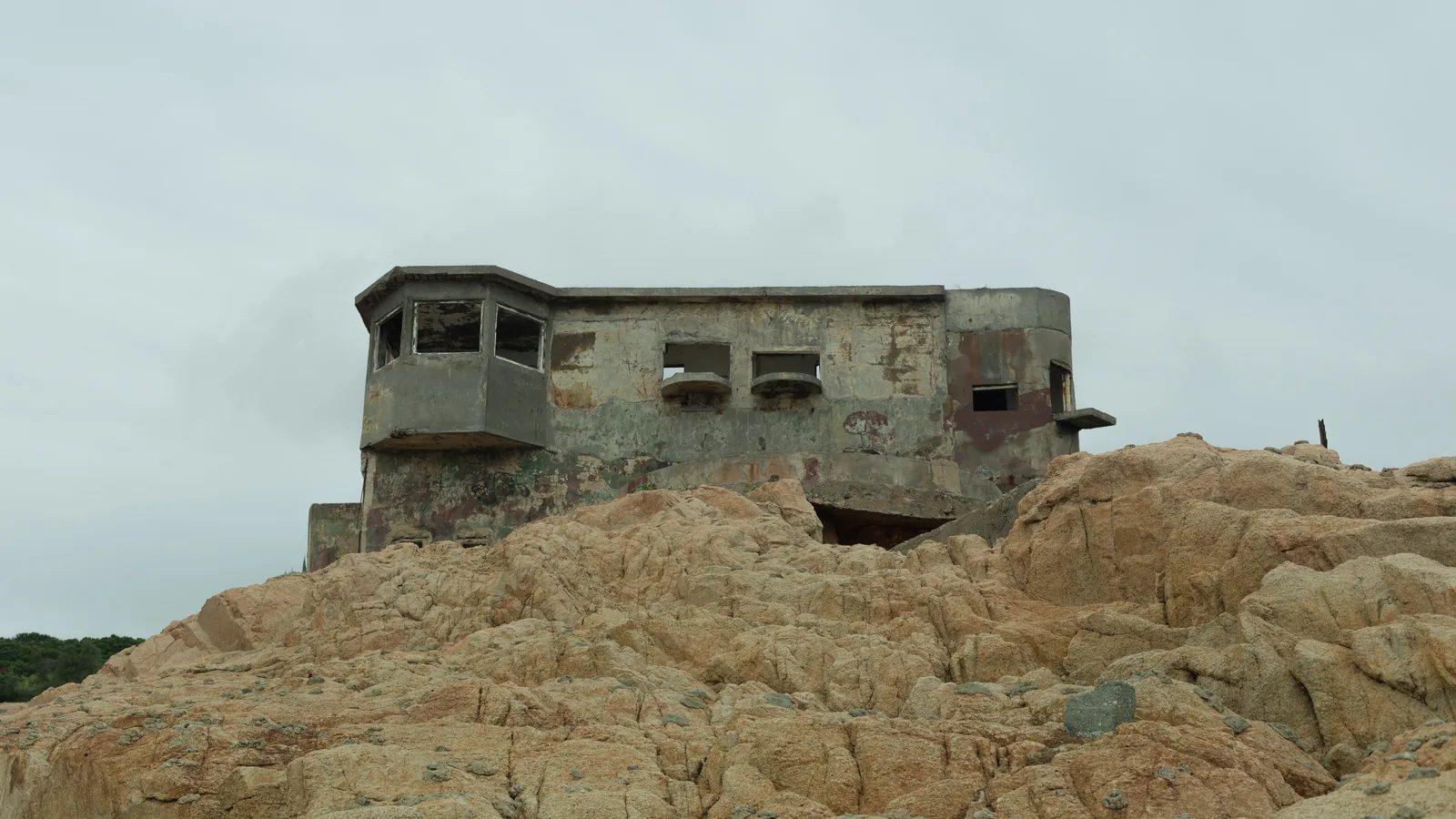 一座外牆斑駁、帶有多個窗洞的混凝土建物坐落在淺褐色岩石上，背景是灰白天空。