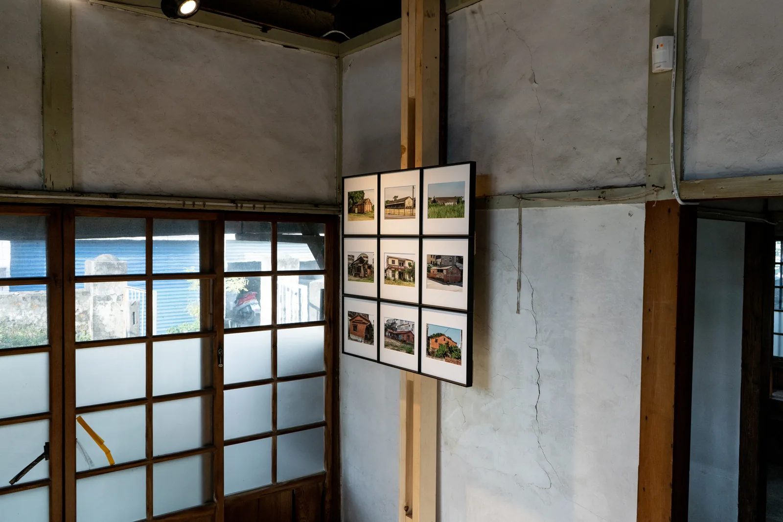 九幅建築影像組成的作品掛在木柱與灰白牆面前，一旁是大片格窗。